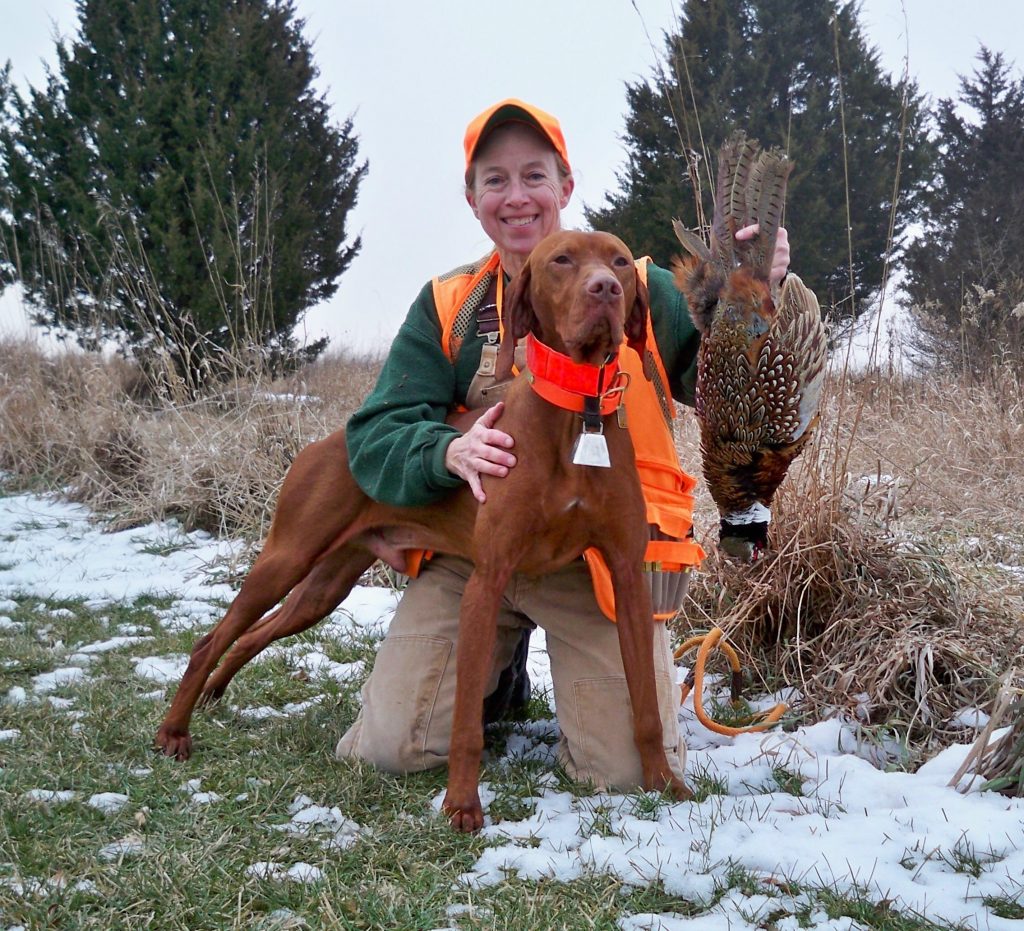 A hunter kneeling next to her hunting dog displays a successful pheasant harvest.