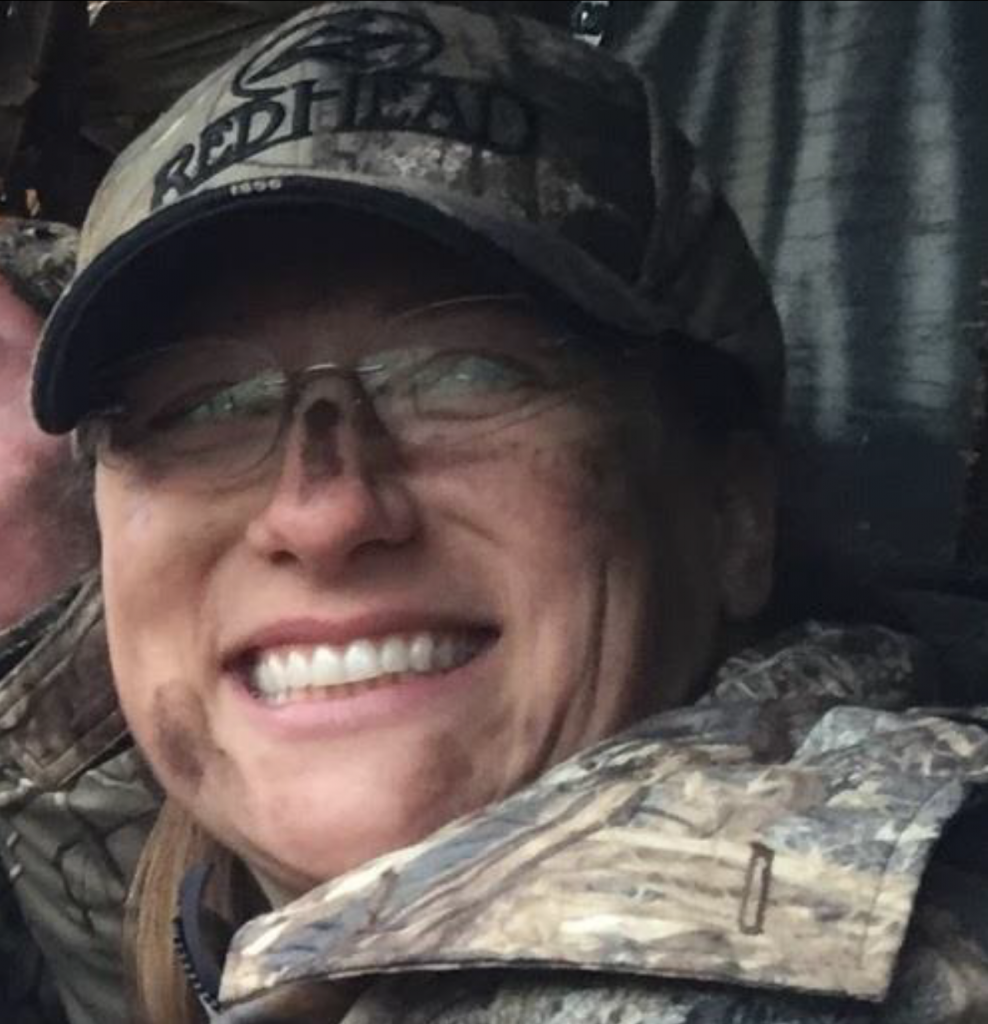 A close-up portrait of a smiling young woman wearing camouflage gear and a camouflage hat.
