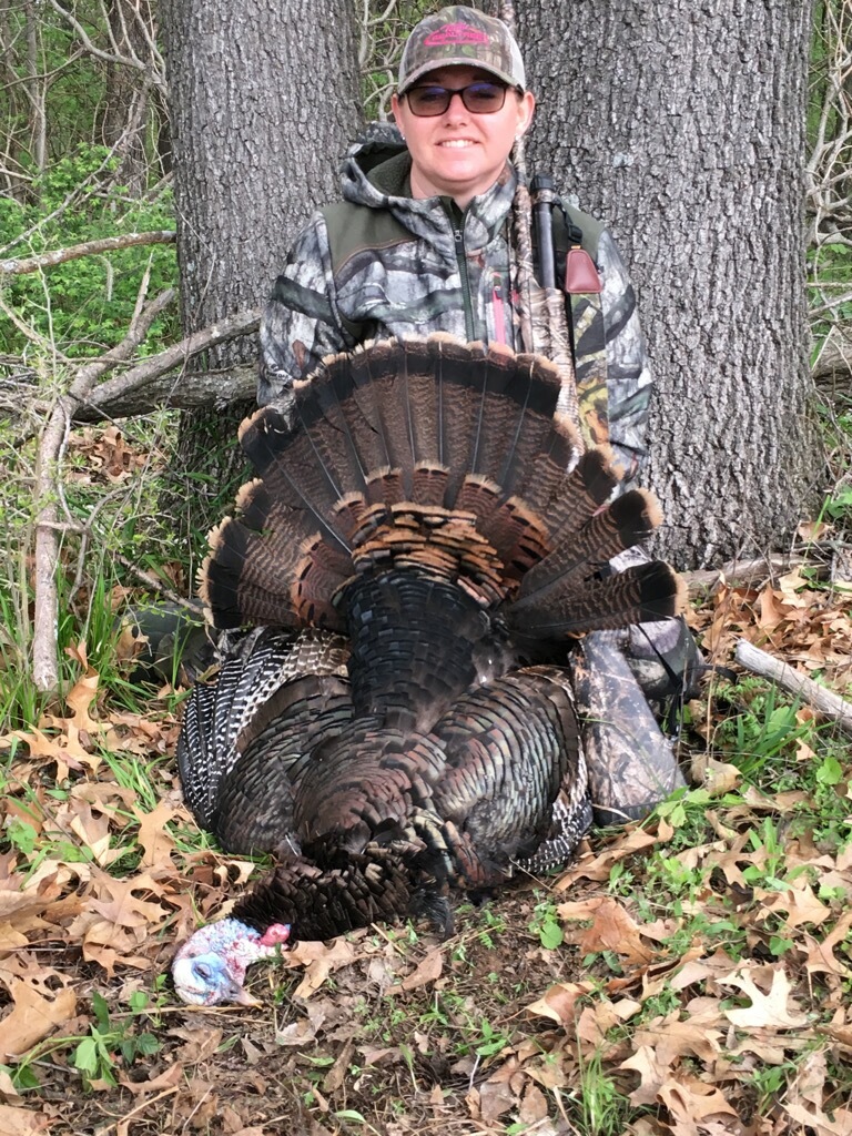 A hunter kneels behind her successfully harvested wild turkey and spreads the tail feathers in a fan. In the background is a tree and a brushy area.