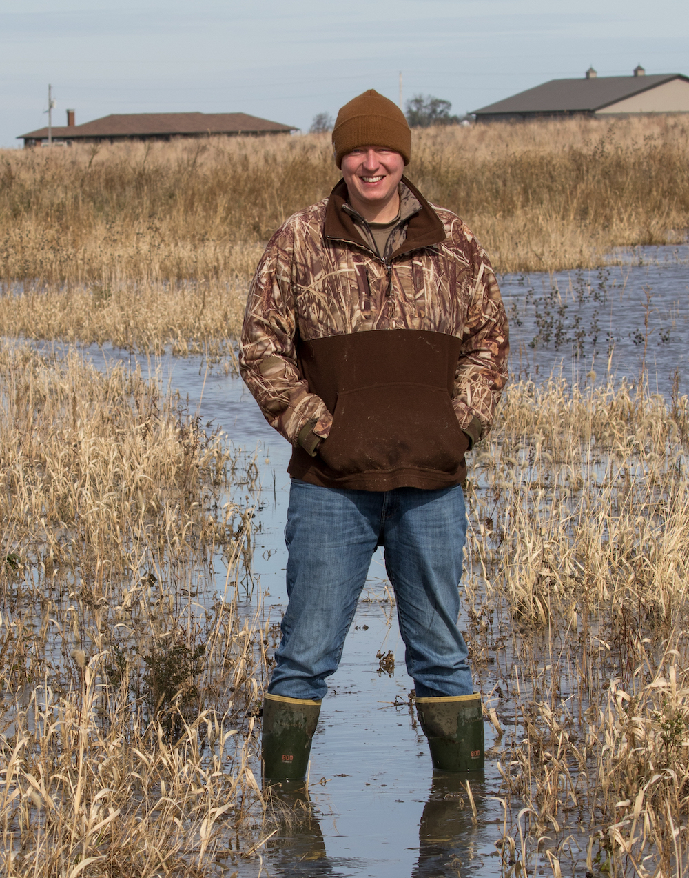 A portrait of a biologist wearing a camouflage jacket, denim jeans, and a tan knitted winter cap. The biologist is standing in a wetland. In the background is tan grasses and vegetation and the roofs of two buildings.