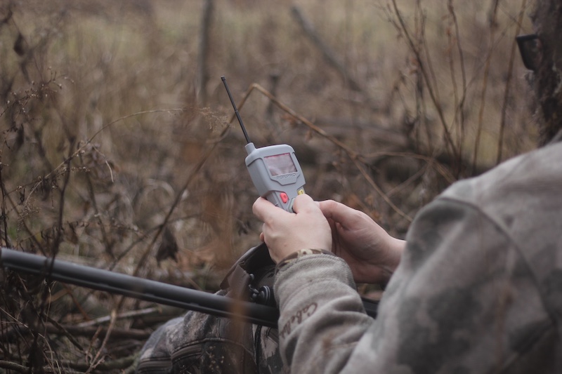 A hunter in camouflage gear sits on the edge of a grassland. The hunter holds a small device in his hands. The device is gray with buttons, a screen, and an antenna. The hunter has a firearm resting in his lap.