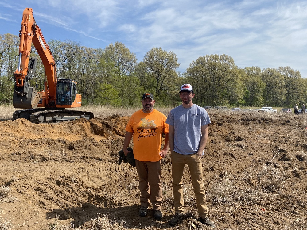 Two men stand in an area cleared out by a backhoe as part of a project to create a new wetland. Both are wearing t-shirts and ballcaps. There are seven vehicles and a treeline in the background of the image.