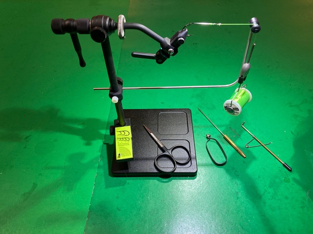 On a green tabletop sits a black metal vice with a rectangular base holding a fishing hook tied to green thread. There is a pair of black scissors resting on the base of the vice as well.