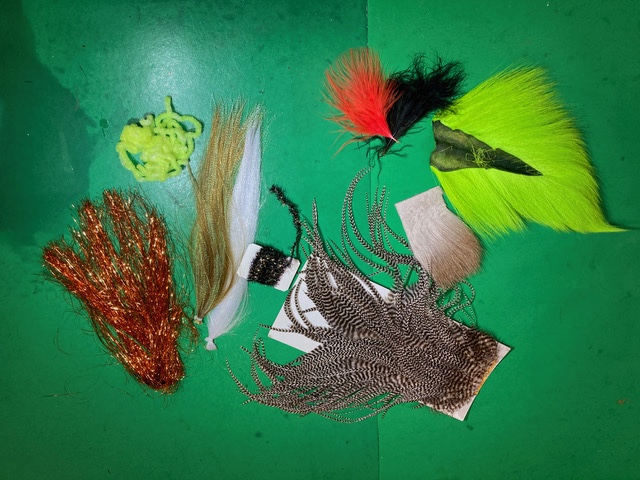 On a green tabletop sits an assortment of natural and dyed feathers, fur, and synthetic fibers.