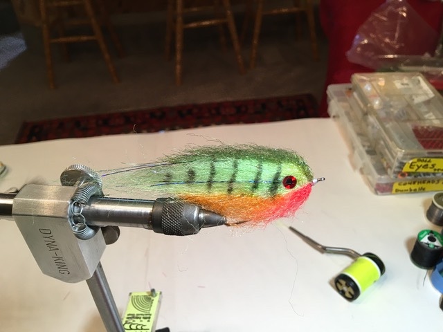 A green and red-orange fishing lure resembling a small fish made of feathers is held by its hook in a metal vice.