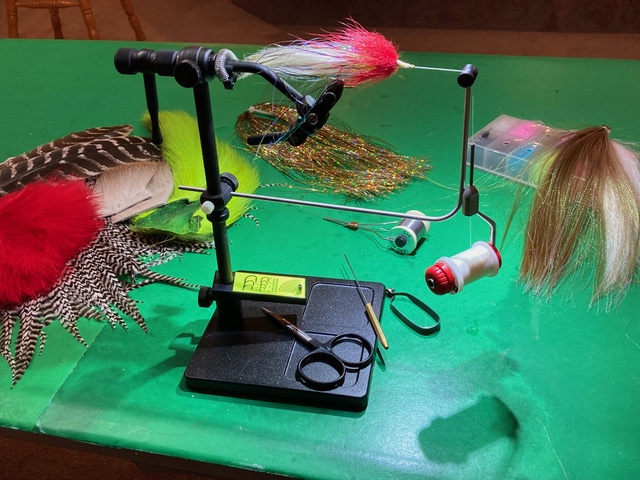 On a green tabletop a black metal vice holds a red and white fishing lure made of different types of fur or fibers by its hook. In the background an assortment of natural and dyed feathers, fur, and synthetic fibers.